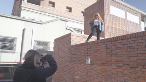 two friends have fun taking pictures in the street. redhead model poses on top of a brick wall and smiles for her photographer friend