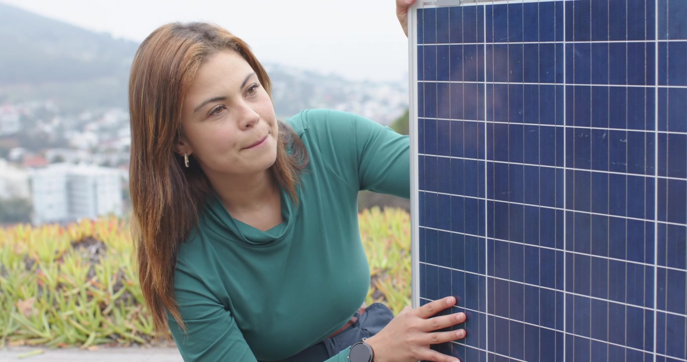 Donna afroamericana inginocchiata con un top color verde acqua che stabilizza un pannello solare e ne controlla l'allineamento sul tetto.