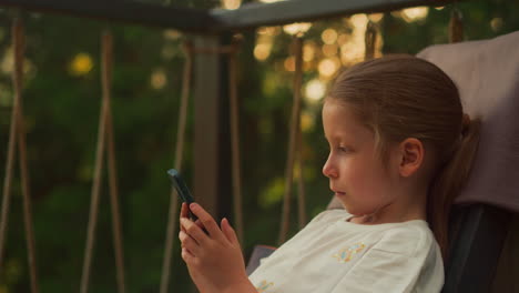 young girl types messages holding smartphone on terrace. toddler relaxes in cozy armchair surrounded by woodland area. calm child rests alone at sunset