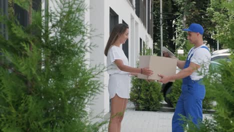 beautiful woman meets a delivery man who gives her a parcel box beside her home, she signs electronic signature on tablet. concept of courier, home delivery, online shopping