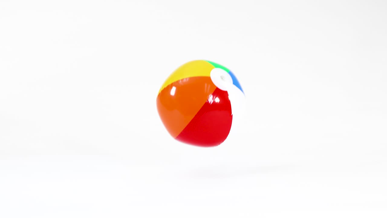 A multi-colored inflatable beach ball bounces and rolls across a bright white studio background