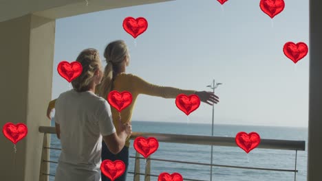 Animation-of-social-media-red-heart-balloons-over-couple-in-love-on-balcony-by-seaside