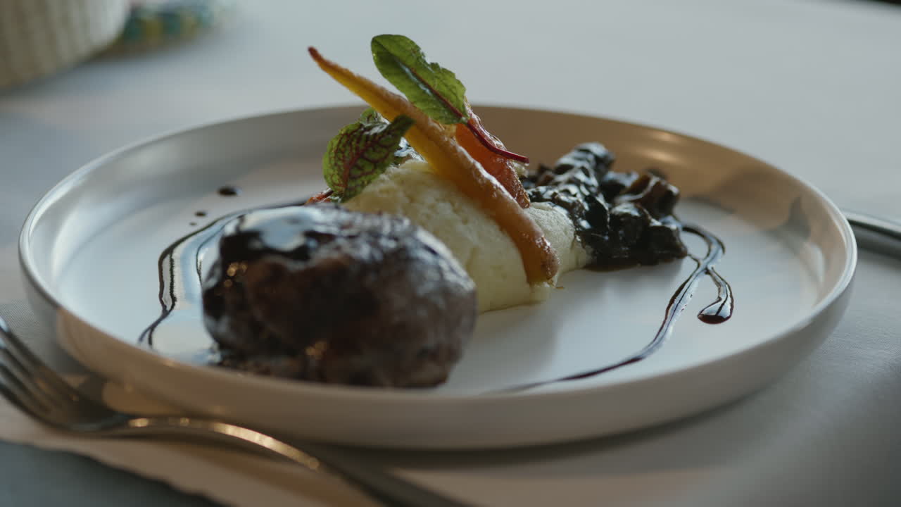 Close-up of a waiter serving a gourmet dish with mashed potatoes, baby carrots, sprouts, and a piece of meat with dark gravy