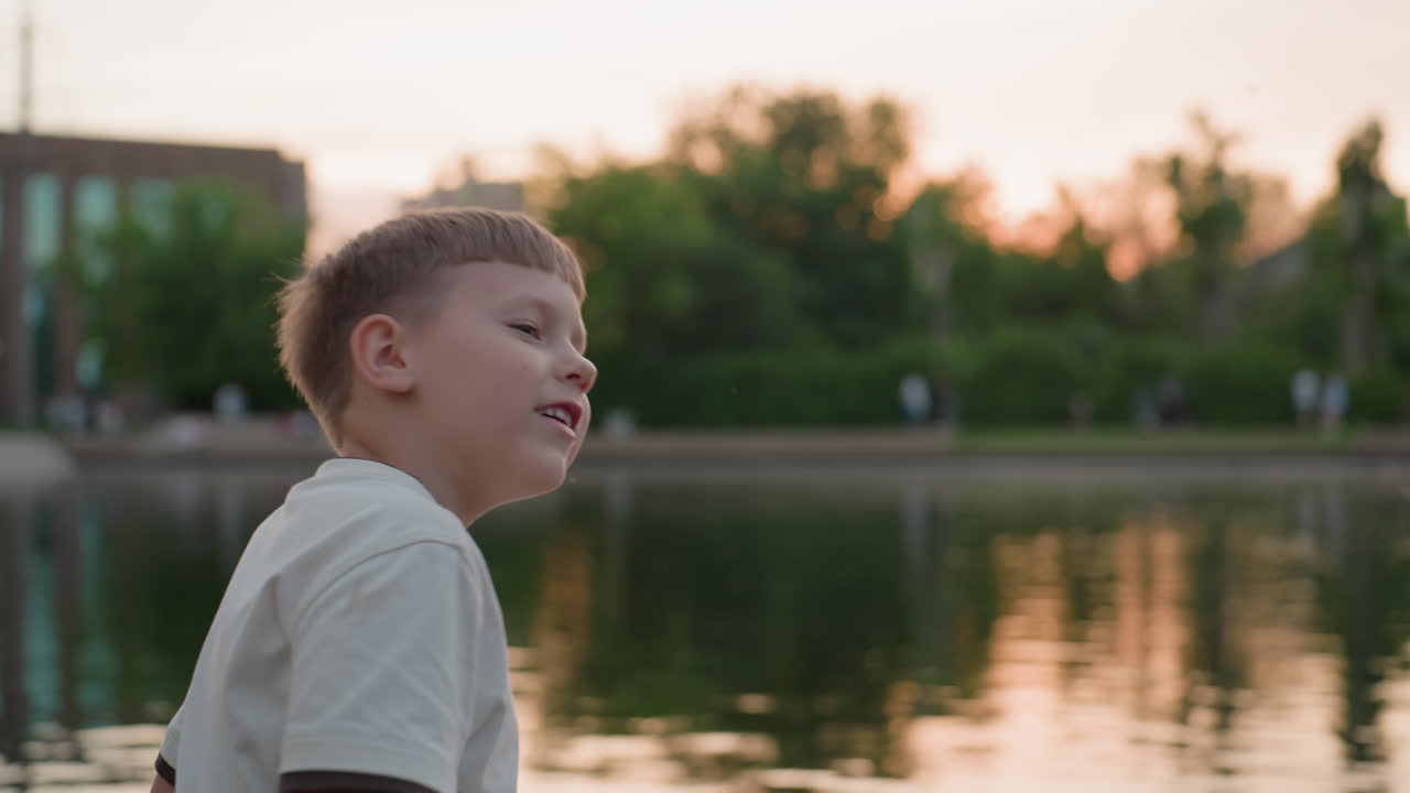 playful boy seated on wooden bridge across calm river at sunset touching water reflection, outdoors calm evening scene with warm golden light and urban park backdrop soft breeze