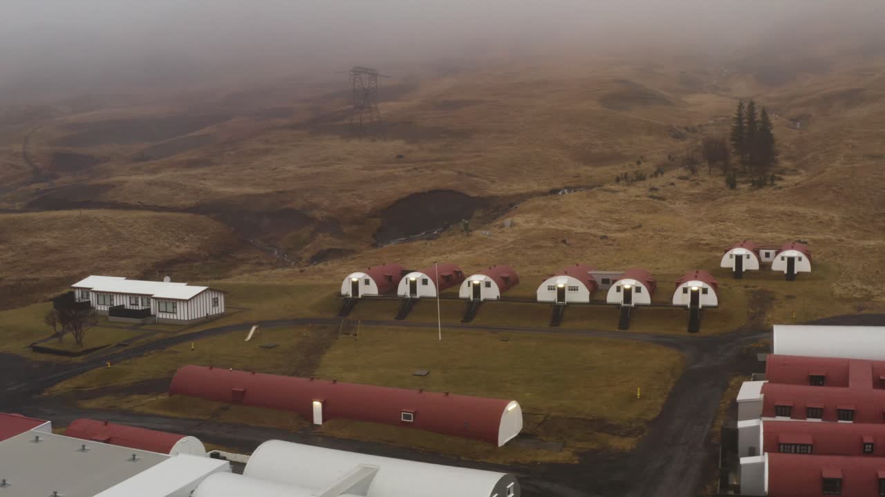 Aerial drone view of red and white Quonset huts aligned on green lawns in Hvalfjörður Iceland with foggy hills and rural landscape in the background during overcast weather