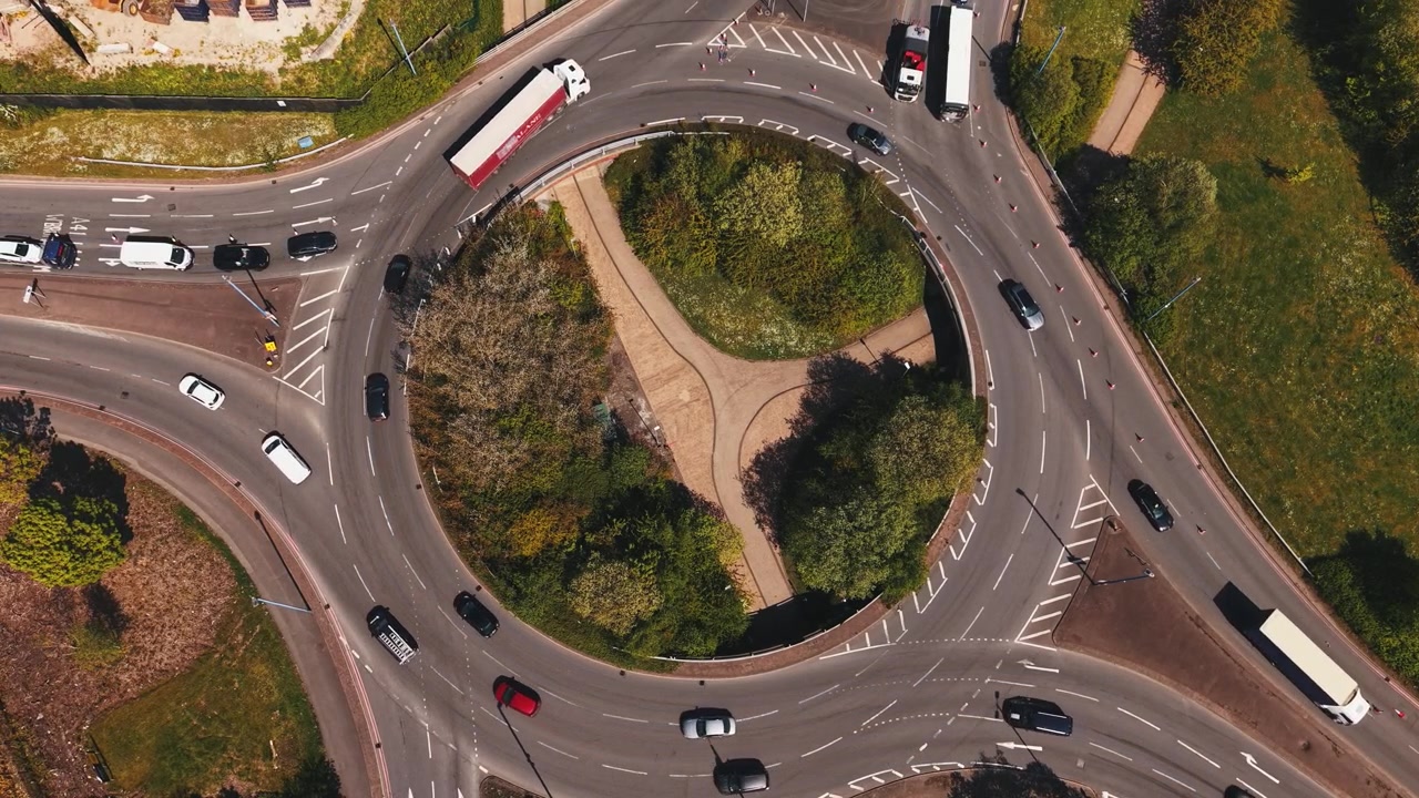 Top down drone footage of cars and trucks moving through busy roundabout in England. Aerial urban traffic scene showing transportation flow and road infrastructure.