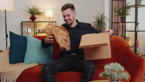 Happy-young-guy-shopper-unpacking-cardboard-box-delivery-parcel-online-shopping-purchase-at-home