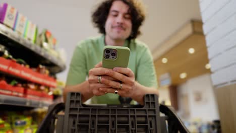 Curly-brunette-guy-uses-his-green-smartphone-while-texting-while-shopping-in-a-supermarket