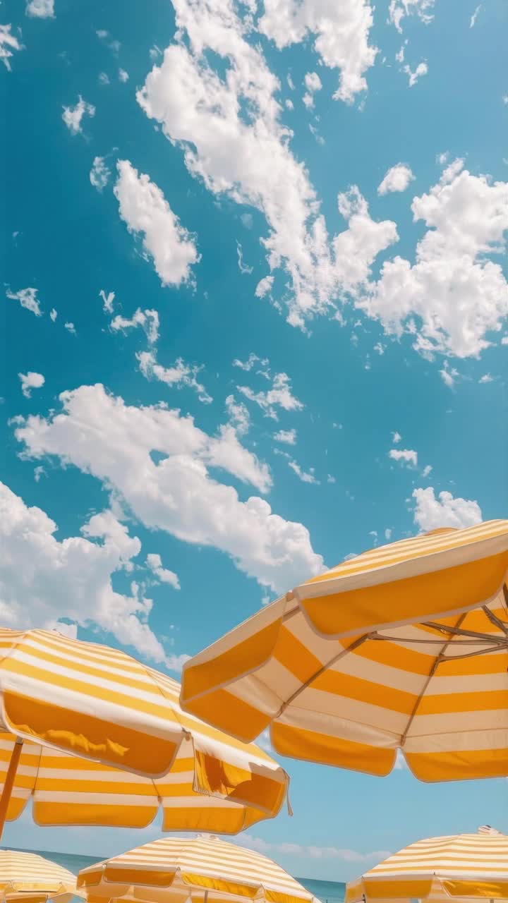 Low-angle video frame of yellow-striped beach umbrellas against a bright blue sky with scattered