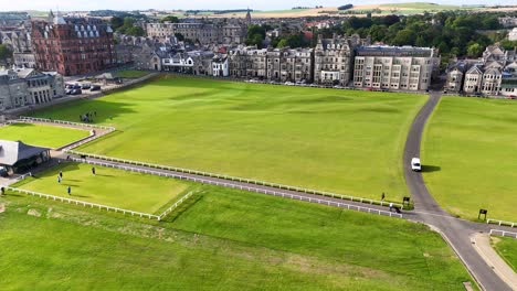 drone footage glides over a lush green golf course bordered by victorian brick buildings in st andrews, scotland, under bright daylight