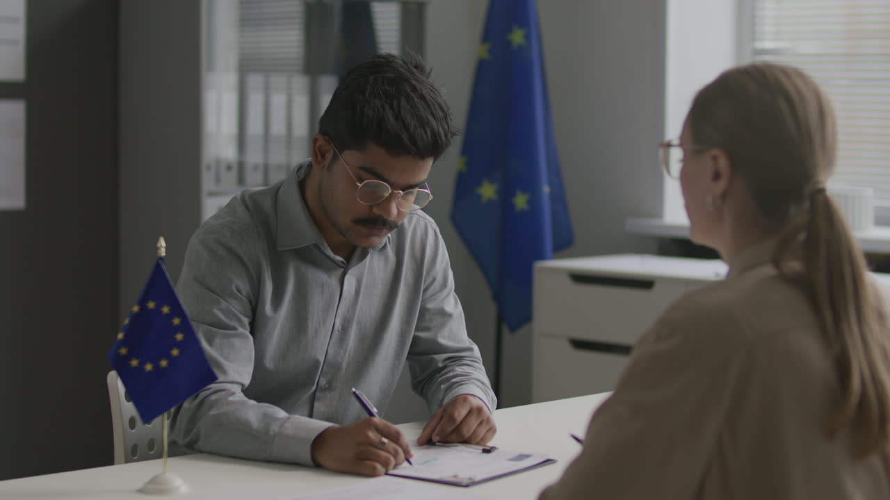 Indian Man Signing EU Visa Application Form under Guidance of Consular Officer