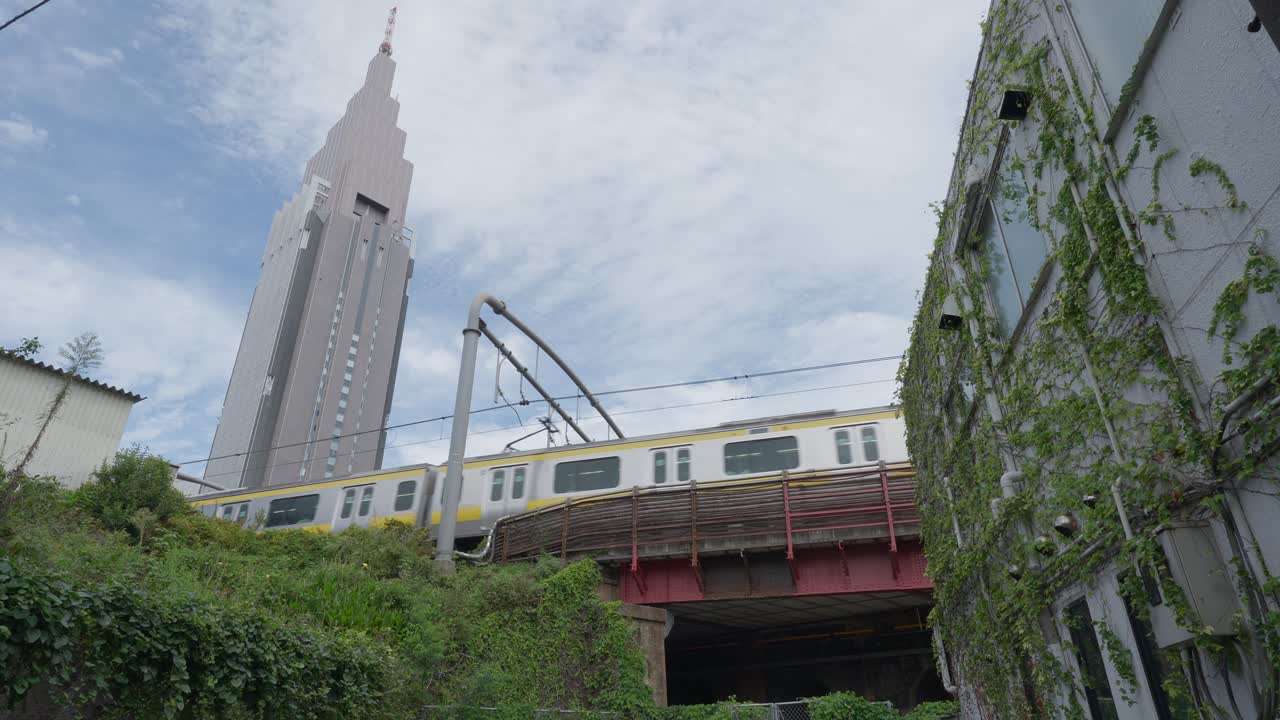 A low-angle shot of a yellow JR commuter train passing over an elevated track, with the towering Docomo Tower in the background