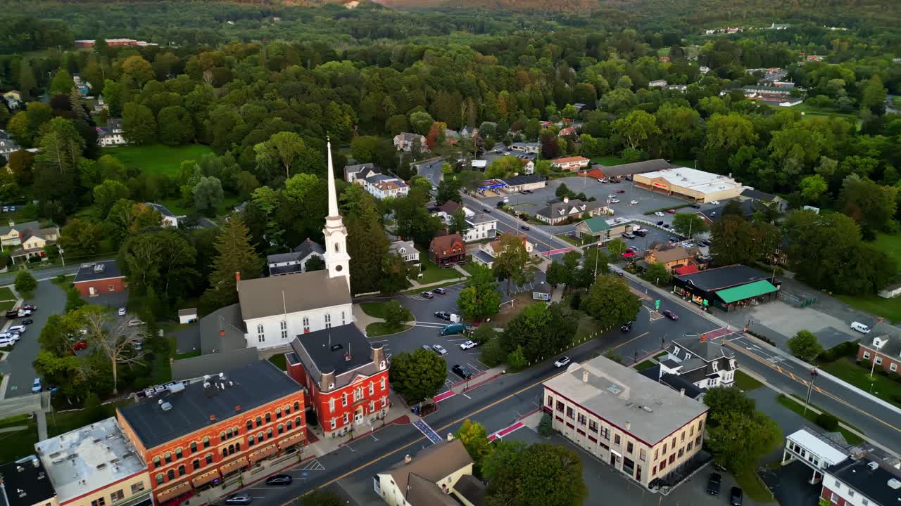 Row of buildings in different colored facade in downtown of american city. Dusk scene in Lee, MA. Colored trees in fall season. Church tower and parking areas. Aerial wide shot. Flyover