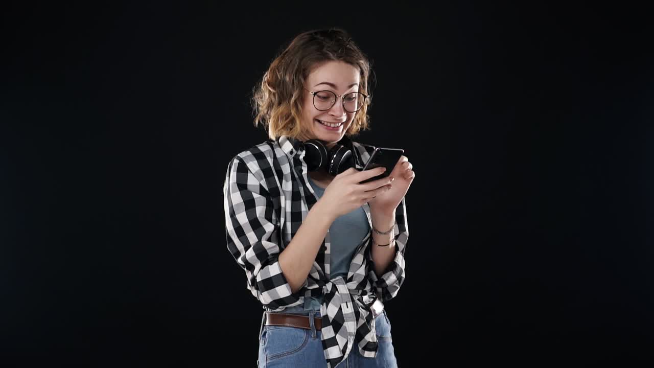 mujer alegre con ropa casual - camisa a cuadros y vaqueros, sosteniendo un teléfono inteligente y desplazándose por los medios sociales o charlando, aislada sobre un fondo negro. emocionándose con las noticias que lee