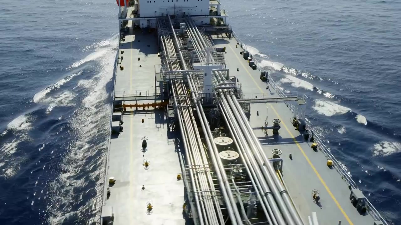 High-angle view follows industrial vessel underway across sunlit water, revealing long metal pipe runs, open deck fittings, red hull sides, foaming wake, and shifting overhead perspective.