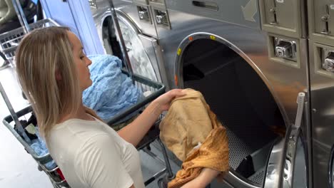 blonde woman putting laundry in commercial dryer at laundromat