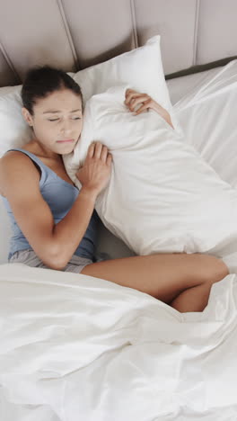 Vertical video of sad biracial woman lying in bed holding pillow at home, slow motion