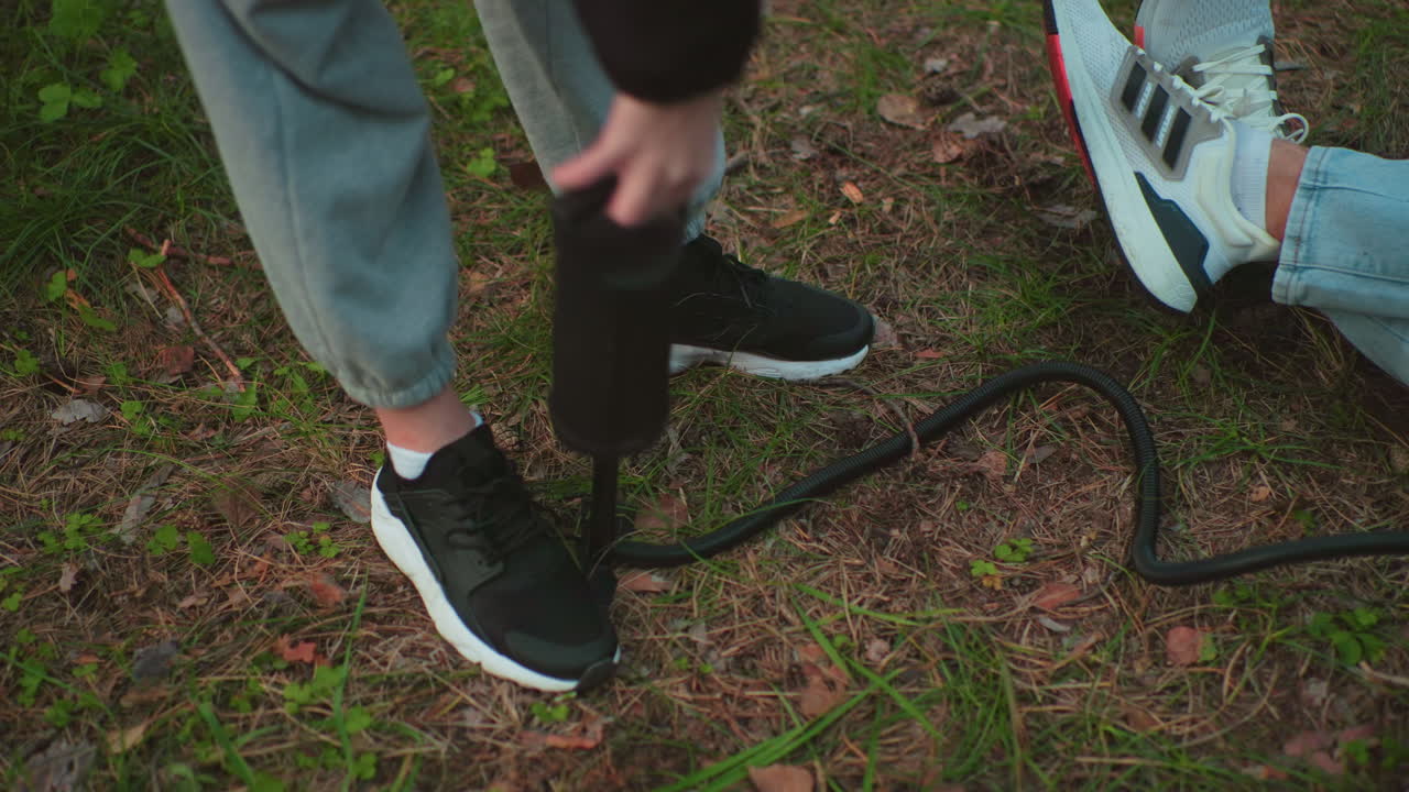 close up of person in joggers and sneakers pressing hand pump into ground while inflating object as another person wearing jeans and white sneakers lays on canvas surface in grassy forest area