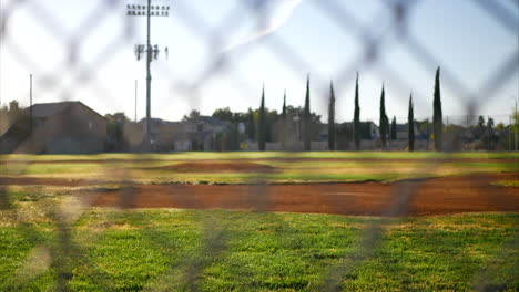 Slide-down-shot-looking-out-at-an-empty-green-baseball-field-diamond-from-behind-home-plate-and-a-chain-link-fence-in-a-local-park-at-sunrise