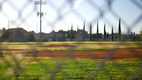tiro deslizado hacia abajo mirando un campo de béisbol verde vacío desde detrás del plato de home y una cerca de eslabones de cadena en un parque local al amanecer