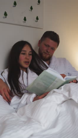 man caresses loose hair of wife reading book in bed. relaxed couple in soft bathrobes enjoys reading novel in bedroom at night. romantic atmosphere