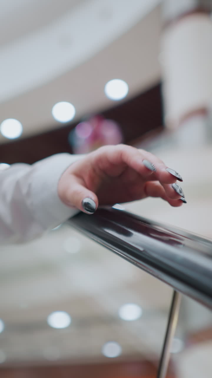 검은색 은 손을 가진 여자의 부분적 인 시각은 하강하는 동안 에스컬레이터 드레일을 부드럽게 니다. 그녀는 색 블라우스와 청바지를 입고 있습니다. 흐릿한 배경은 현대 쇼핑몰을 포착합니다.