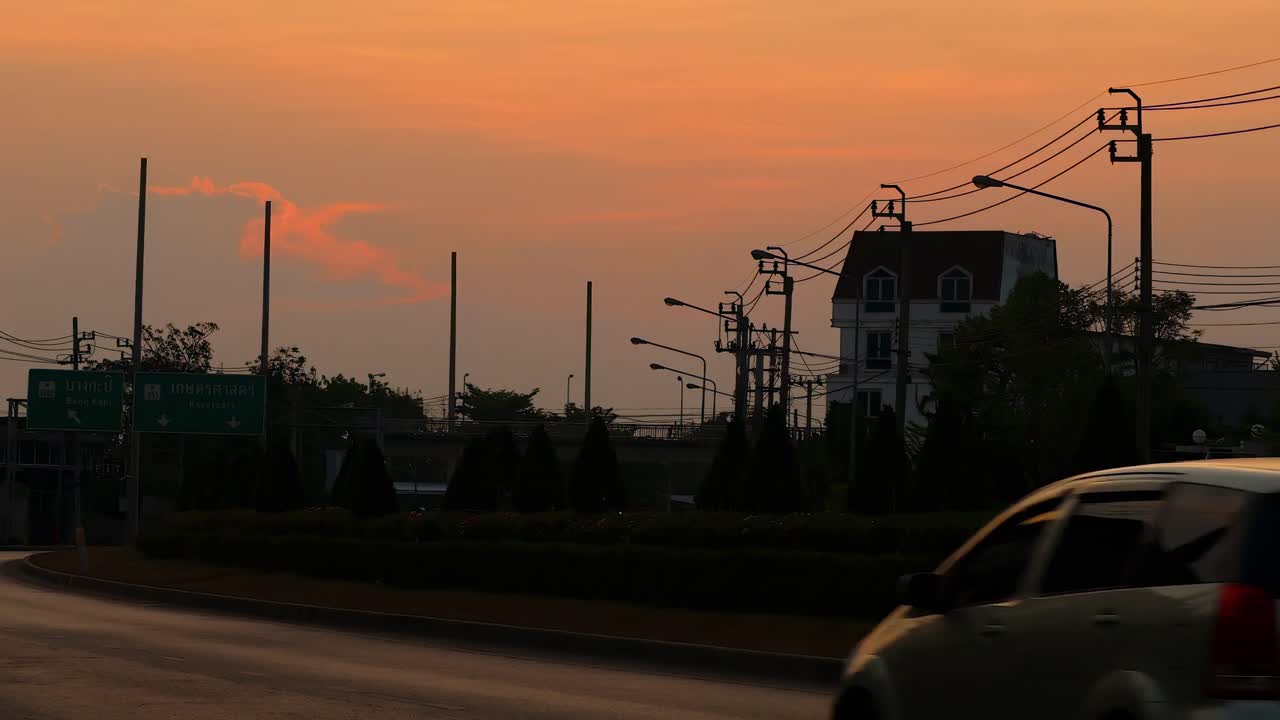 Cars drive along a road as the sun sets, casting an orange glow over the urban landscape.
