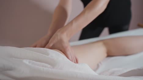 massage therapist in black using thumbs to deeply massage client thigh while client lies under white sheet in softly lit therapy room with gentle shadows and relaxed setting