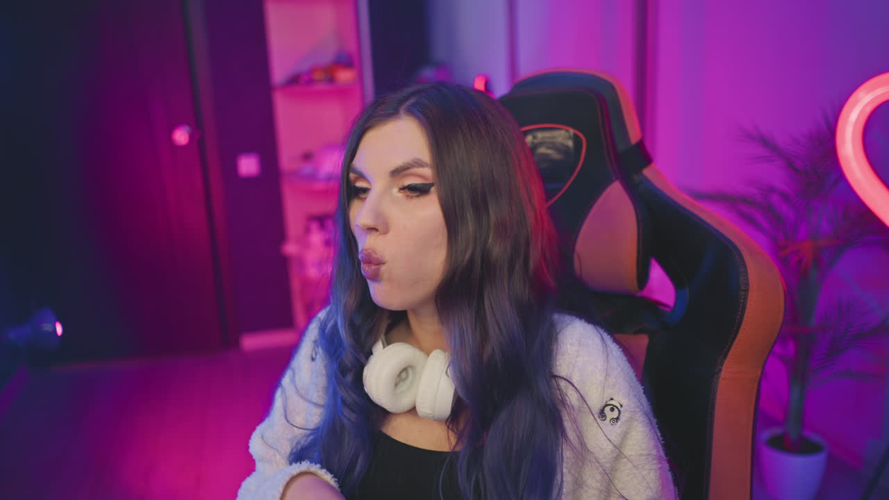 Fair skinned lady with dark hair seated in gaming chair eating popcorn, eyes fixed intently off camera, wearing headphones around neck against vibrant pink and purple neon lit studio backdrop