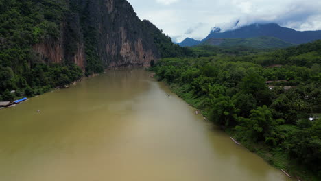 Eine-Drohne-Bewegt-Sich-über-Den-Fließenden-Mekong-In-Der-Nähe-Von-Pak-Ou-In-Laos-Mit-Wolkenbedeckten-Bergen-In-Der-Ferne