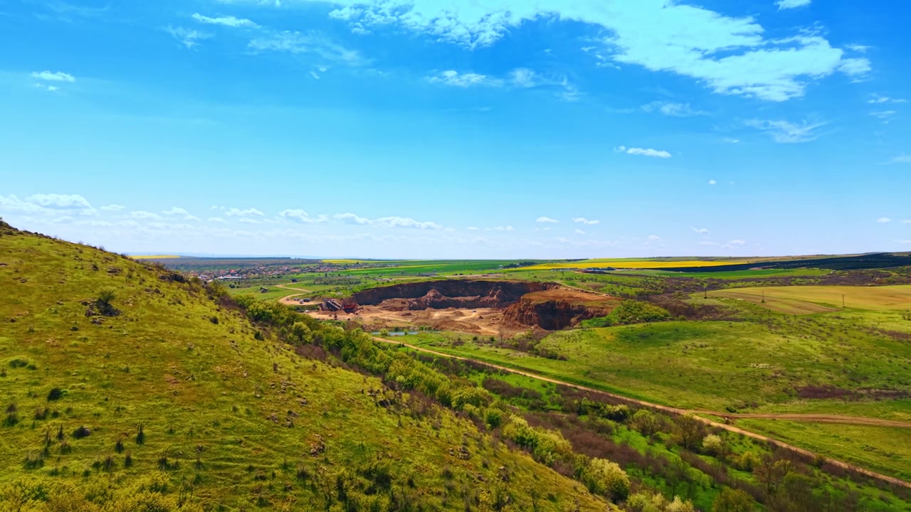 Fugleperspektiv av et dagbrudd omgitt av grønne åser og jorder. Et vidt landskap viser et utgravd steinbrudd blant grønne gresskledde skråninger og jordbruksland under blå himmel.