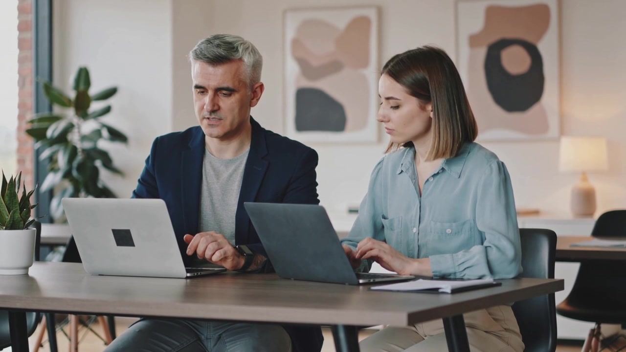 Uomo in blazer che digita, mentre una donna osserva, digita e controlla il lavoro a un tavolo d'ufficio con laptop