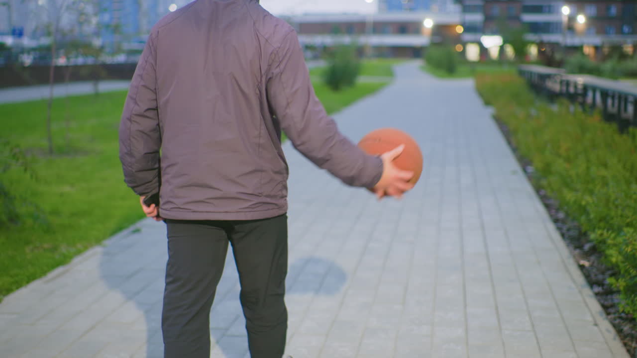 evening urban walk holding basketball, unspecified race person seen from back in jacket and jeans, strolling along paved park path flanked by grass and railing, soft bokeh city lights, cinematic
