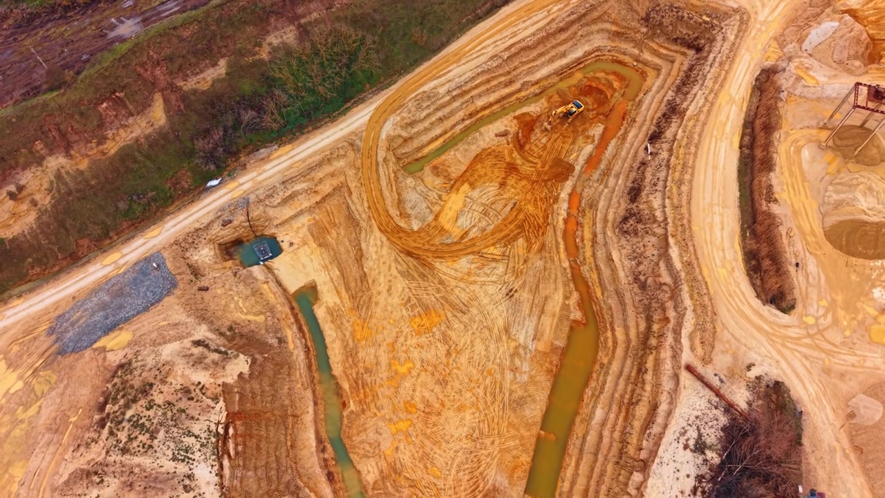 Aerial view of open pit sand quarry with heavy equipment. Overhead drone perspective of excavator, water channels, gravel piles and forest edge.