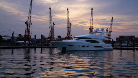 white luxury yacht moored in a serene harbor, surrounded by industrial cranes in the background, creating a striking contrast between opulence and industry during a stunning sunset
