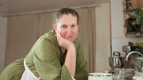white lady in green dress and white apron gently rests head on hand while leaning on kitchen counter, looking at camera with warm soft smile, surrounded by baking items