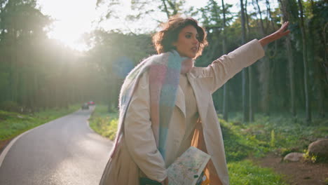 travelling girl catching car on road sunlit forest holding map. woman tourist