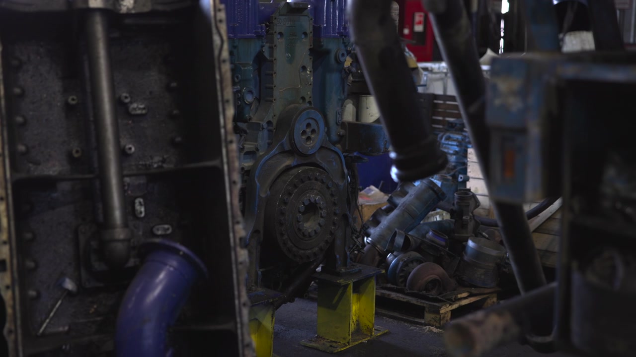 Fokus i mellemgrunden på en stor adskilt blå motorblok med et blotlagt cirkulært svinghjul monteret på gule metalstativer inde i et dæmpet industriværksted, med mørke skygger, slidte overflader og reparation