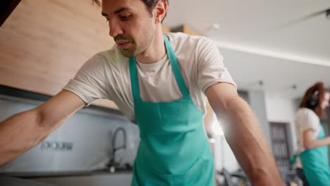 an inside view from the camera a brunette guy with stubble in a white t-shirt and a blue apron washes the stove in a modern kitchen while using a cleaning company