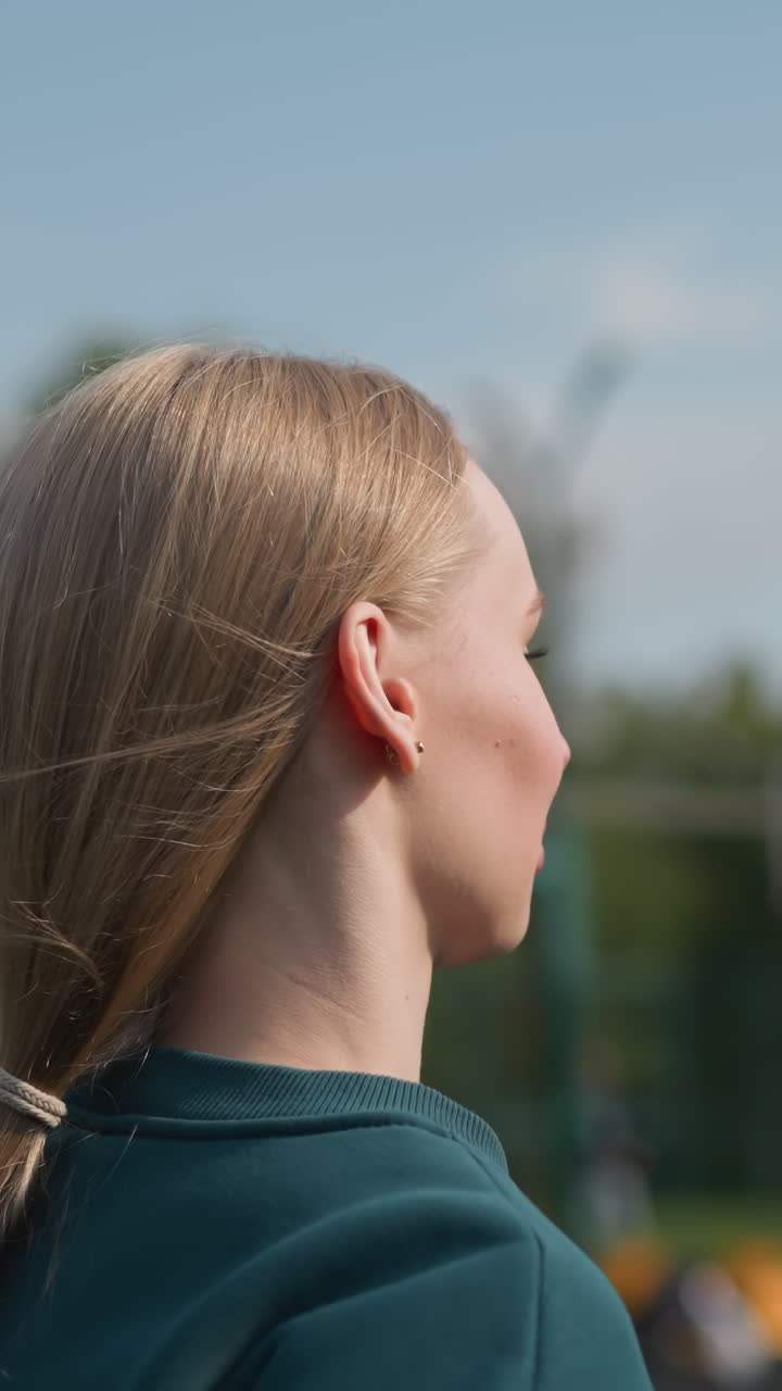 Primo piano di una donna con i capelli raccolti e un maglione verde che fa jogging in un campo da beach volley all'aperto con rete da pallavolo e alberi sullo sfondo, catturando il movimento atletico durante l'allenamento fitness