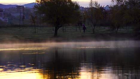 water lake steaming at morning on city park, people walking around trees