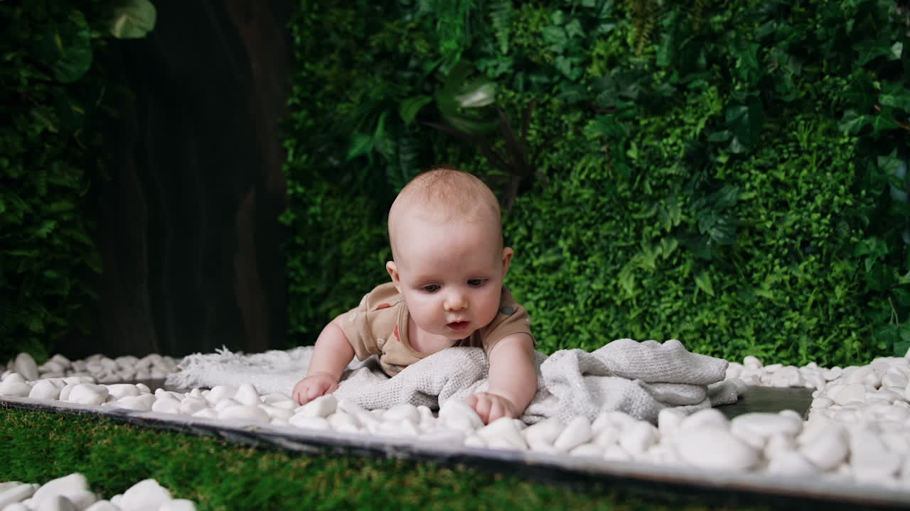 Beautiful infant baby lies outdoors on the plaid. Little infant is interested with white pebbles around him. Greenery at backdrop.