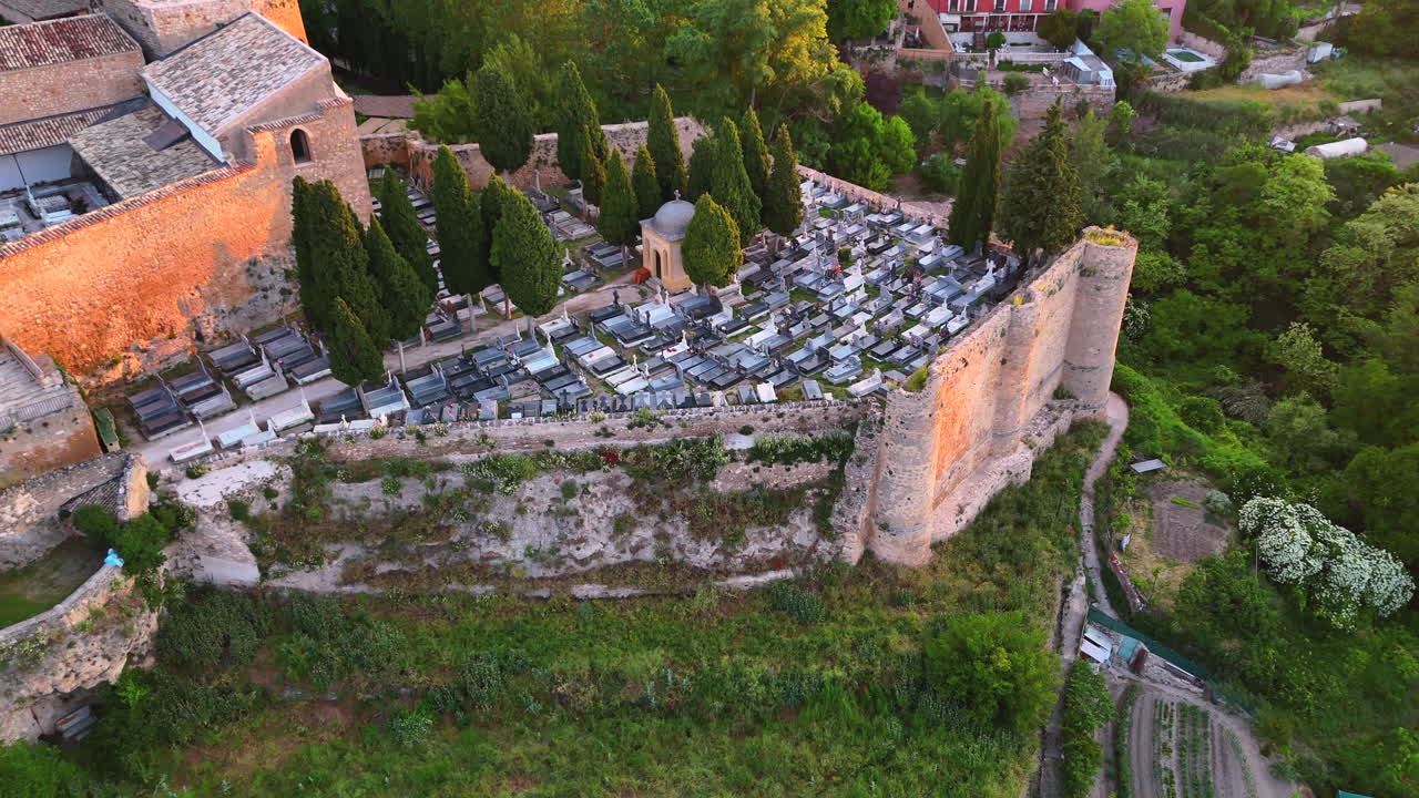Drone flies backward and upward over the historic walled cemetery in Brihuega, La Alcarria, Spain. The scene reveals gravestones, trees, chapel and ancient walls in warm sunrise light