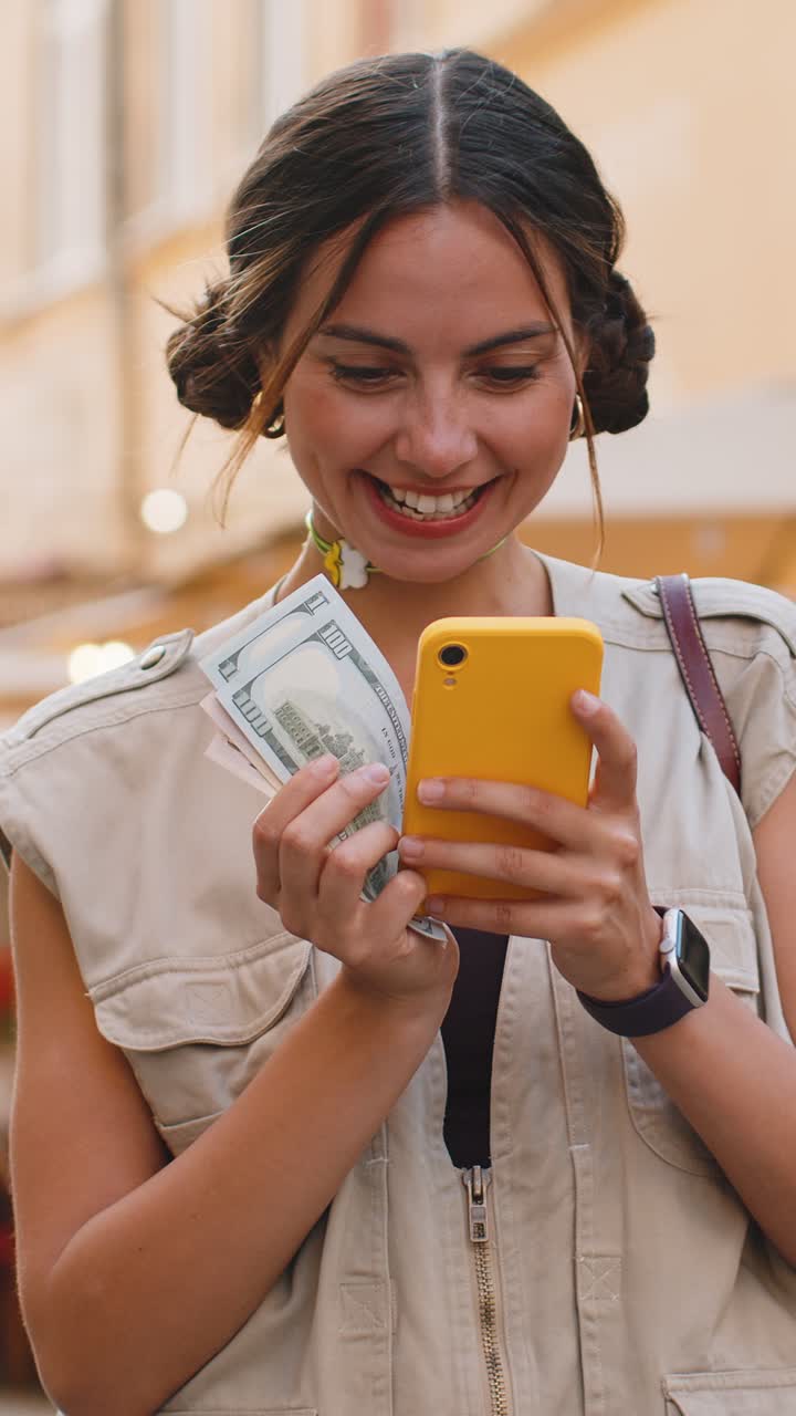 Giovane donna felice che conta soldi, dollari in contanti, usa un'app calcolatrice per smartphone in una strada urbana.