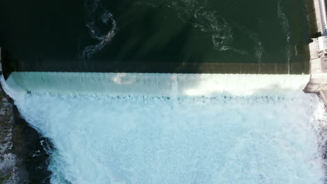 An overhead shot of a wide dam shows the split between still water and roaring falls. Structures line both sides of the river