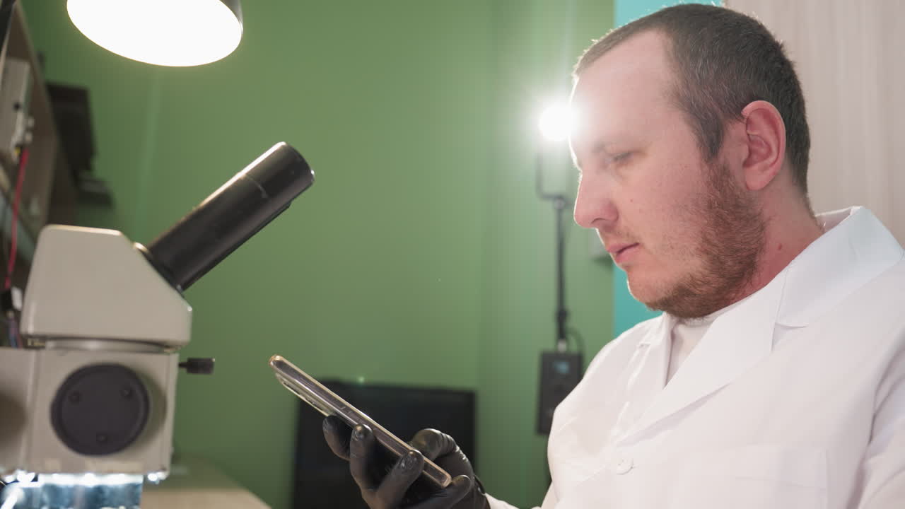 백색 실험실 코트와 검은 장갑을 입은 기술자의 근접 뷰, 실험실에서 현미경 아래 회로를 조사하는 동안 전화로 이야기하고, 책상 램프를 켜고