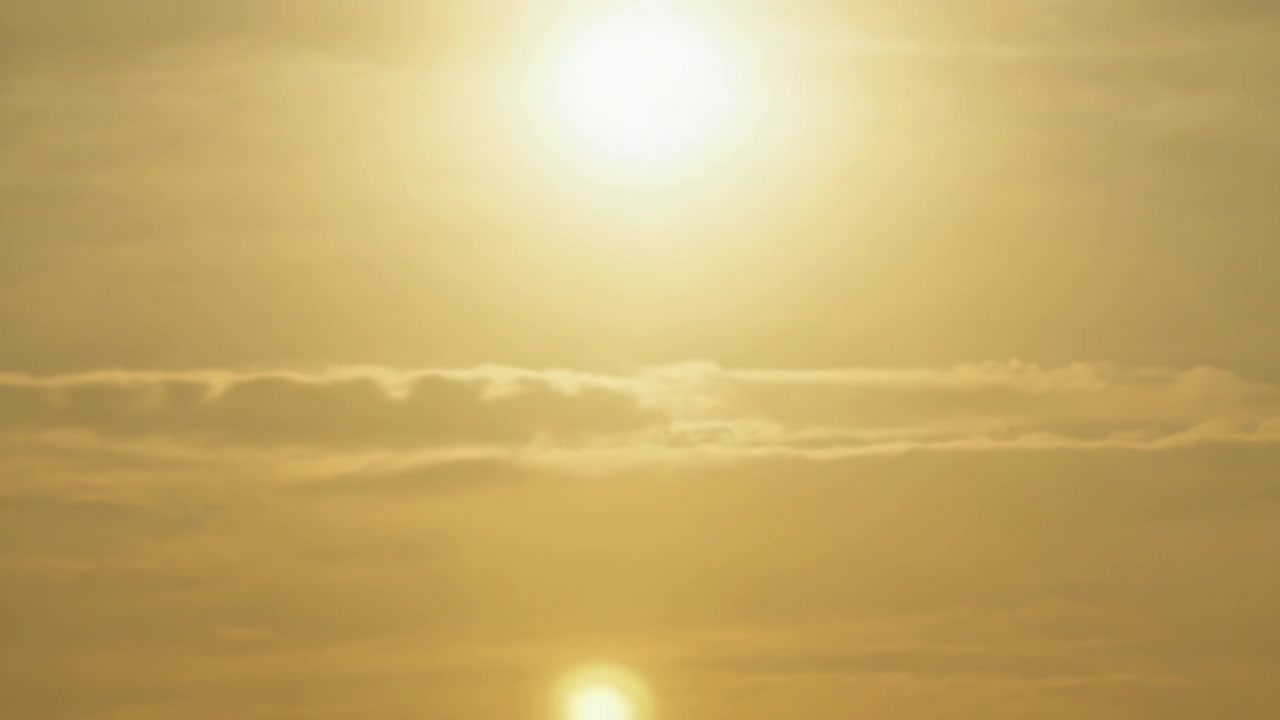 Lysende sol og tiltagende lysskær nedadtil over kysthorisonten med diset varm himmel og skybånd.