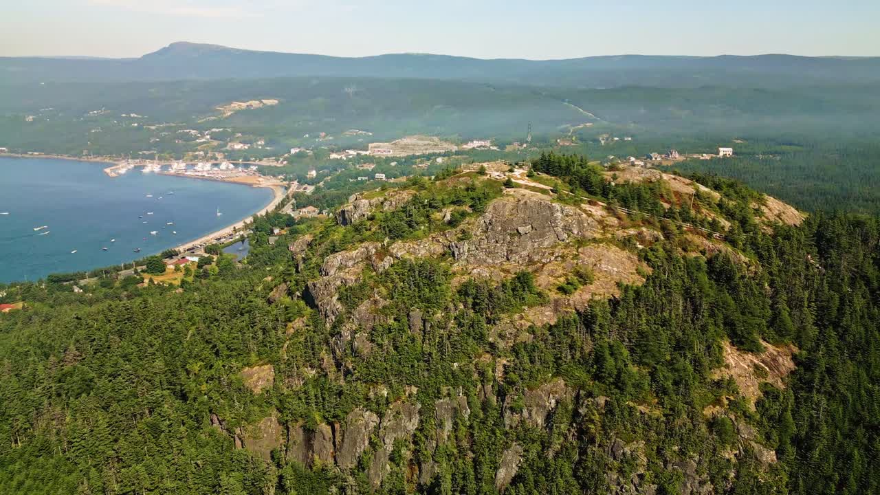 A drone soars over Holyrood's lush hills and bay as a generating station emits a plume of smoke rising into the clear sky, contrasting with the surrounding green forests and water