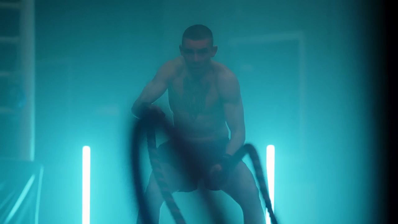 Kraftfuld mand træner med battle ropes i et mørkt, blåtbelyst fitnesscenter
