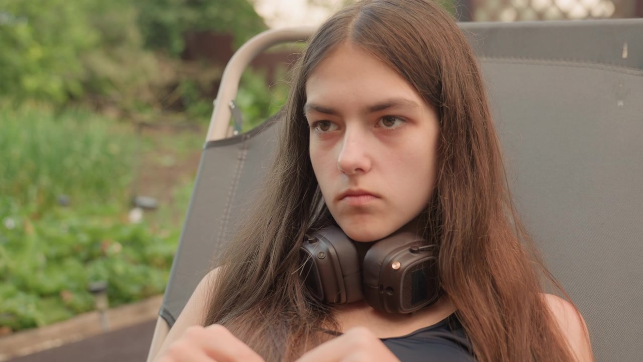 Una giovane donna con lunghi capelli castani siede all'aperto su una sedia grigia, con cuffie nere al collo e che sistema un oggetto tra le mani. Lo sfondo presenta vegetazione ed elementi da giardino